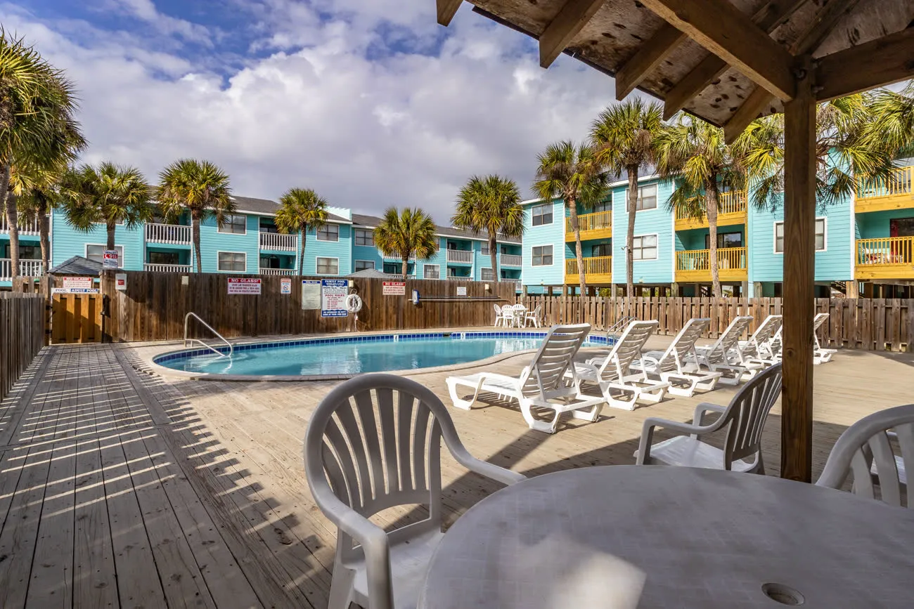 Adults pool with chairs and table foreground, palm trees behind